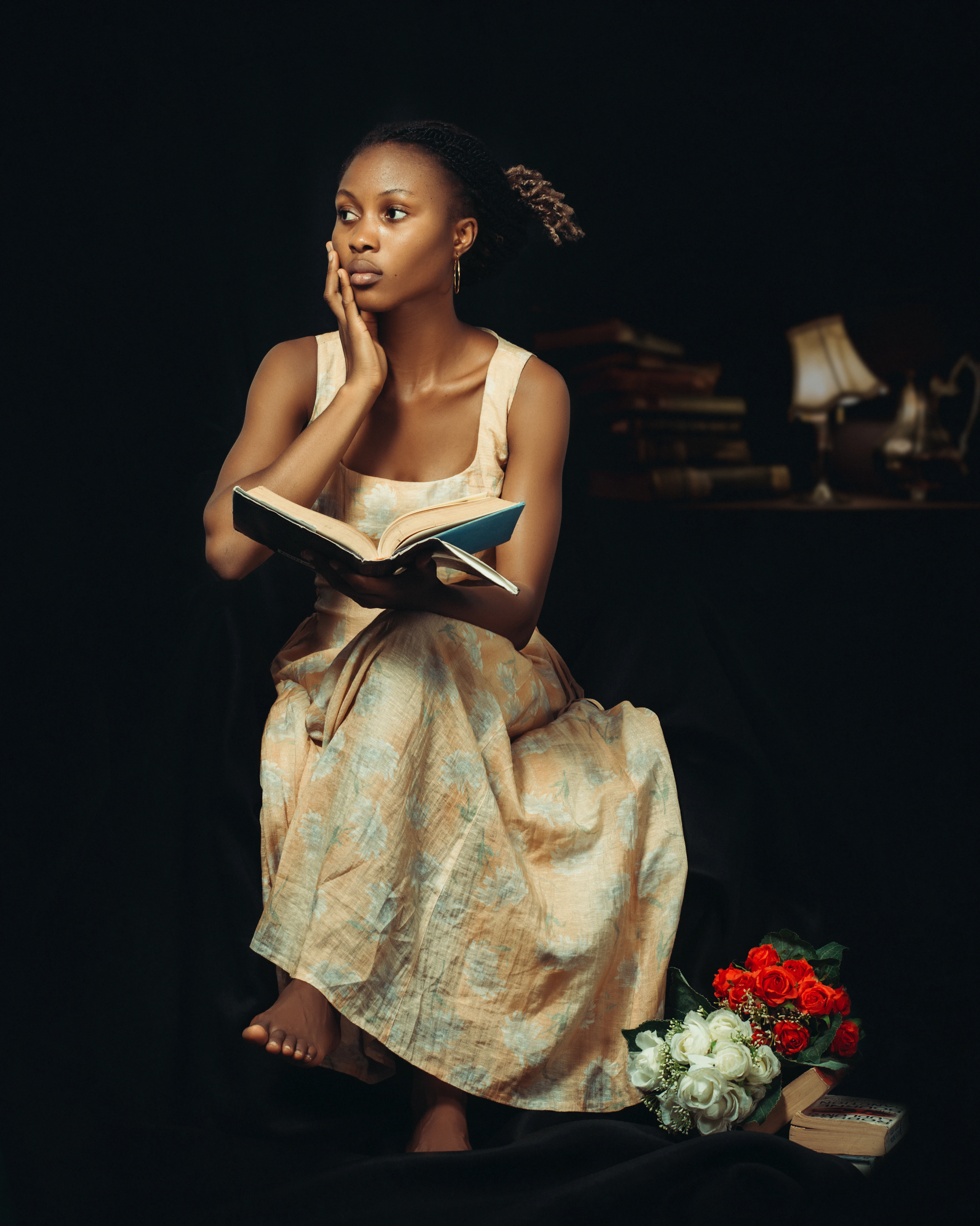 A woman in a cream dress sitting and reading an open book against a dark backdrop