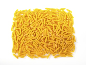 A large pile of uncooked yellow penne pasta is scattered against a plain white background. The cylindrical, tube-shaped pasta pieces are neatly arranged, showcasing their smooth texture and hollow centers.