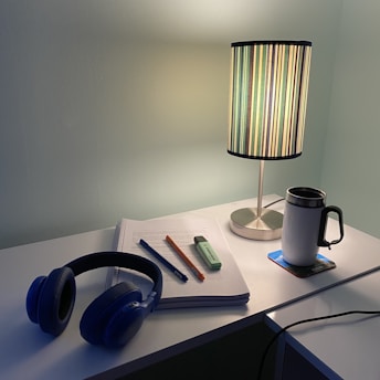 A desk with a study setup including a notebook, a blue pen, an orange mechanical pencil, a green highlighter, and black headphones. A table lamp with a striped shade provides warm light. A white travel mug sits on a coaster beside the notebook.