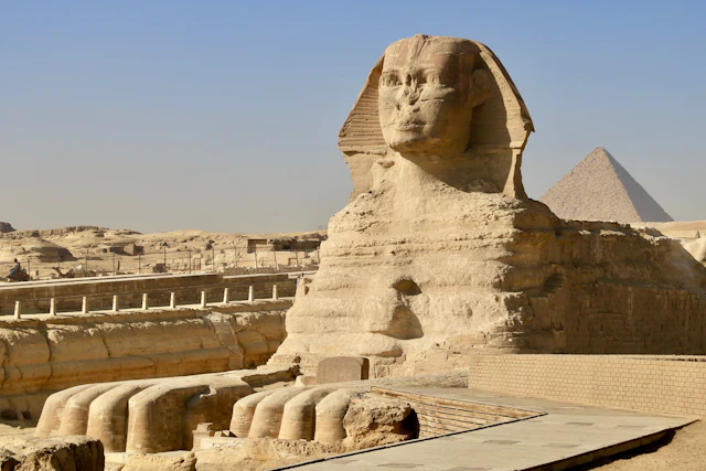 The Great Sphinx of Giza in profile with one of the pyramids visible behind it against a bright blue sky.