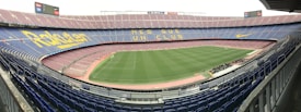 A large football stadium with empty stands and a well-maintained pitch in the center. The seating is arranged in tiers around the field, with blue and maroon seats. Advertising banners and slogans are displayed prominently, and the phrase 'MES QUE UN CLUB' is visible across the seats.