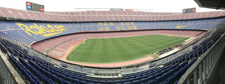 A large football stadium with empty stands and a well-maintained pitch in the center. The seating is arranged in tiers around the field, with blue and maroon seats. Advertising banners and slogans are displayed prominently, and the phrase 'MES QUE UN CLUB' is visible across the seats.
