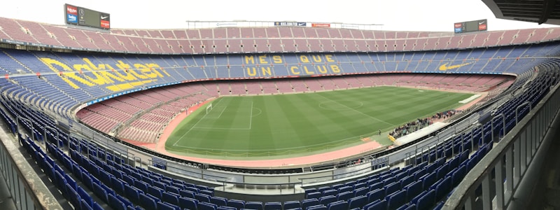 A large football stadium with empty stands and a well-maintained pitch in the center. The seating is arranged in tiers around the field, with blue and maroon seats. Advertising banners and slogans are displayed prominently, and the phrase 'MES QUE UN CLUB' is visible across the seats.