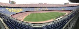 A large football stadium with empty stands and a well-maintained pitch in the center. The seating is arranged in tiers around the field, with blue and maroon seats. Advertising banners and slogans are displayed prominently, and the phrase 'MES QUE UN CLUB' is visible across the seats.