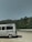 A white van with a roof rack and a graphic of trees and a tent on its side is parked on a gravel area. In the background, there is a landscape of green trees and hills under a clear sky.