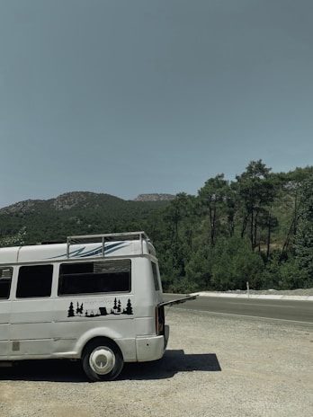 A white van with a roof rack and a graphic of trees and a tent on its side is parked on a gravel area. In the background, there is a landscape of green trees and hills under a clear sky.