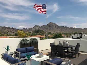 A rooftop patio with modern outdoor furniture and an American flag flying against a backdrop of mountains. The patio has blue cushioned chairs and small green plants in pots, with a table set that includes wicker chairs and decorative vases.