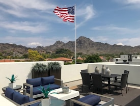 A rooftop patio with modern outdoor furniture and an American flag flying against a backdrop of mountains. The patio has blue cushioned chairs and small green plants in pots, with a table set that includes wicker chairs and decorative vases.