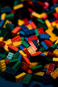 A pile of colorful interlocking plastic bricks scattered haphazardly. The pieces are primarily red, yellow, blue, and green, each featuring multiple studs on top for connecting with other pieces. The chaotic arrangement of the blocks creates a vibrant and playful appearance.