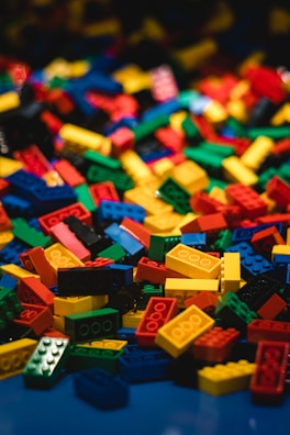 A large pile of interlocking building blocks in various shapes and sizes is scattered across a surface. The blocks are primarily bright primary colors, standing out vividly against a dark background.