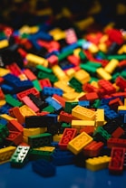 A large pile of interlocking building blocks in various shapes and sizes is scattered across a surface. The blocks are primarily bright primary colors, standing out vividly against a dark background.