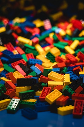A large pile of interlocking building blocks in various shapes and sizes is scattered across a surface. The blocks are primarily bright primary colors, standing out vividly against a dark background.