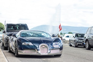 A luxurious sports car with sleek, aerodynamic design is parked on the street, surrounded by other vehicles. The scenery suggests an urban area with a backdrop of distant mountains and a water fountain. A Swiss flag is prominently displayed nearby, hinting at a possible location.