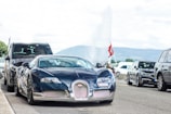 A luxurious sports car with sleek, aerodynamic design is parked on the street, surrounded by other vehicles. The scenery suggests an urban area with a backdrop of distant mountains and a water fountain. A Swiss flag is prominently displayed nearby, hinting at a possible location.