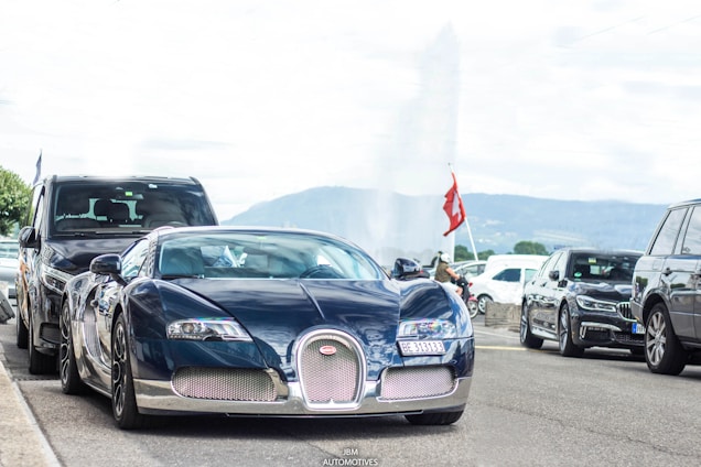 A luxurious sports car with sleek, aerodynamic design is parked on the street, surrounded by other vehicles. The scenery suggests an urban area with a backdrop of distant mountains and a water fountain. A Swiss flag is prominently displayed nearby, hinting at a possible location.
