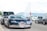 A luxurious sports car with sleek, aerodynamic design is parked on the street, surrounded by other vehicles. The scenery suggests an urban area with a backdrop of distant mountains and a water fountain. A Swiss flag is prominently displayed nearby, hinting at a possible location.