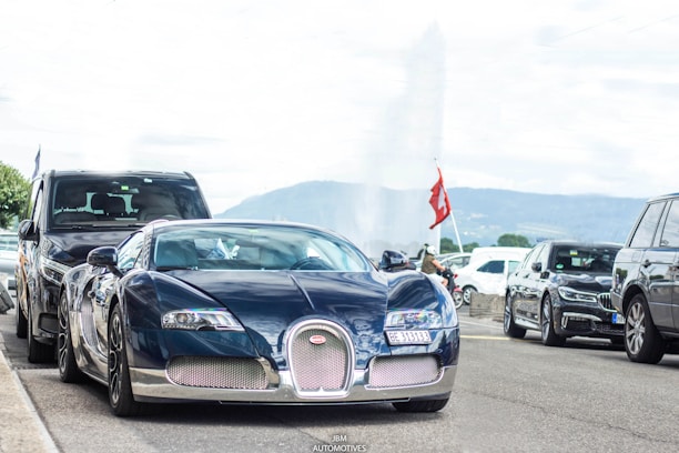 A luxurious sports car with sleek, aerodynamic design is parked on the street, surrounded by other vehicles. The scenery suggests an urban area with a backdrop of distant mountains and a water fountain. A Swiss flag is prominently displayed nearby, hinting at a possible location.
