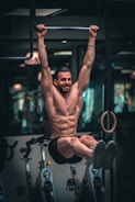 A muscular man performs a hanging leg raise exercise in a gym setting, gripping a pull-up bar with both hands. Behind him, exercise bikes are visible, adding to the fitness environment.