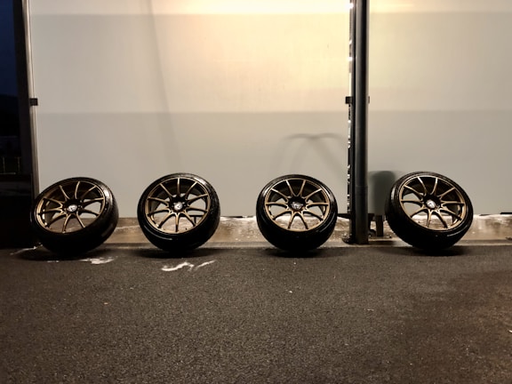 Four car wheels are leaning against a wall in a line on a dark asphalt surface. The wall is light in color with a slight reflection, and there's a black vertical pole on the right side. The lighting creates shadows on the ground.