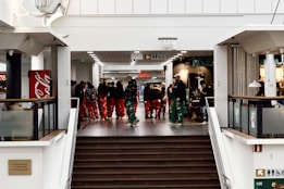 A group of people wearing colorful pants gather in a shopping mall. They are talking and moving through a hallway with various shops and a sale sign in the background. The setting includes a staircase leading upwards and visible branding of a soda company on the left.