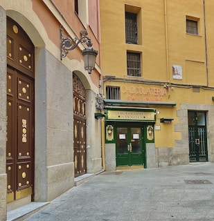 A historic building with a charming facade featuring large wooden doors adorned with gold accents. The shopfront displays a sign for 'Chocolatería San Ginés' in stylish lettering. The walls of the building are painted in soft colors with traditional architectural details, and there is an ornate lamp attached to the wall.