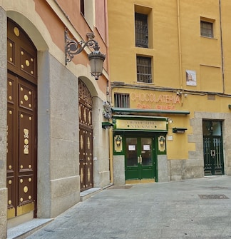 A historic building with a charming facade featuring large wooden doors adorned with gold accents. The shopfront displays a sign for 'Chocolatería San Ginés' in stylish lettering. The walls of the building are painted in soft colors with traditional architectural details, and there is an ornate lamp attached to the wall.