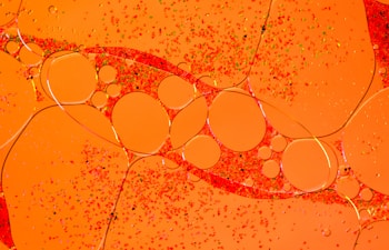A vibrant array of orange and red hues with circular shapes resembling oil droplets floating on a liquid surface. Tiny particles are scattered throughout, adding texture to the smooth, glossy appearance of the larger spheres.