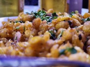 A close-up of a dish filled with golden-brown crispy corn kernels, garnished with fresh green herbs and small pieces of red onion. The focus is on the texture and colors, emphasizing the freshness and crispiness of the food.
