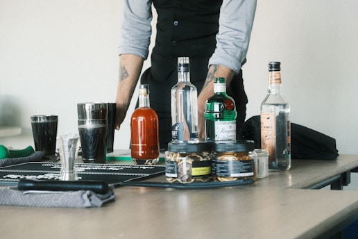 A person stands behind a table with various bottles of alcohol, a cocktail shaker, and glassware. The individual is wearing a light-colored shirt with a dark vest and has tattoos on their forearms. The table is equipped with a bar mat and several ingredients and tools for making cocktails are visible.