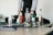 A person stands behind a table with various bottles of alcohol, a cocktail shaker, and glassware. The individual is wearing a light-colored shirt with a dark vest and has tattoos on their forearms. The table is equipped with a bar mat and several ingredients and tools for making cocktails are visible.