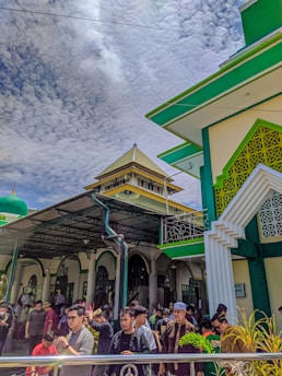 A large group of people gathered outside a mosque, which features distinct green and white Islamic architectural elements. The sky above is partly cloudy, and the scene appears active with various individuals interacting and moving around the entrance.