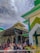 A large group of people gathered outside a mosque, which features distinct green and white Islamic architectural elements. The sky above is partly cloudy, and the scene appears active with various individuals interacting and moving around the entrance.