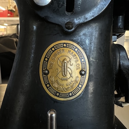 A close-up of an old sewing machine featuring a brass plate with the text 'The Singer Manfg Co' and 'Trade Mark.' The plaque includes an emblem with a threaded needle and decorative elements.