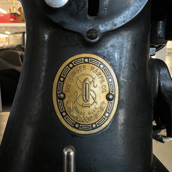 A close-up of an old sewing machine featuring a brass plate with the text 'The Singer Manfg Co' and 'Trade Mark.' The plaque includes an emblem with a threaded needle and decorative elements.