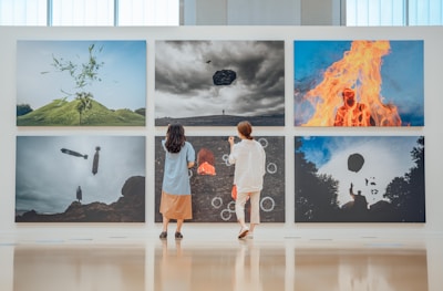 Two individuals are observing an art gallery wall displaying six large photographs. The images feature surreal landscapes and abstract elements, including a floating rock, a fire engulfing a figure, and silhouettes against the sky. The gallery space is bright and minimalist, enhancing the artwork's impact.