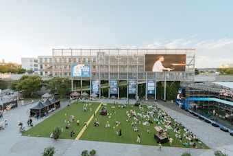 A large outdoor space featuring a green lawn area with numerous people casually sitting, likely enjoying an event. Surrounding the lawn, there are multiple tents and small stalls, some of which display signs with 'World Muyo'. In the background, a modern building with a large digital screen shows a person playing the piano. The setting appears to be in a public venue, possibly a festival or concert space.