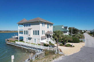 A waterfront house with a light beige exterior, located next to a body of water. The house features multiple levels with large windows and several balconies, surrounded by a white picket fence. The landscape includes palm trees and a small, well-maintained garden. The background shows more houses along a road and a clear blue sky.
