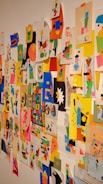 A colorful collage of abstract and geometric art pieces attached to a wall with tape. The images contain a mix of vibrant shapes, characters, and patterns using a variety of colors including pink, blue, yellow, and black. Each piece seems to be individually made using cut-out paper arranged creatively.