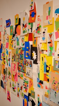 A colorful collage of abstract and geometric art pieces attached to a wall with tape. The images contain a mix of vibrant shapes, characters, and patterns using a variety of colors including pink, blue, yellow, and black. Each piece seems to be individually made using cut-out paper arranged creatively.
