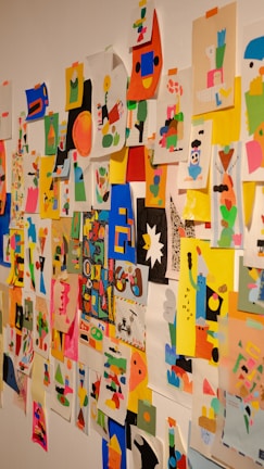 A colorful collage of abstract and geometric art pieces attached to a wall with tape. The images contain a mix of vibrant shapes, characters, and patterns using a variety of colors including pink, blue, yellow, and black. Each piece seems to be individually made using cut-out paper arranged creatively.