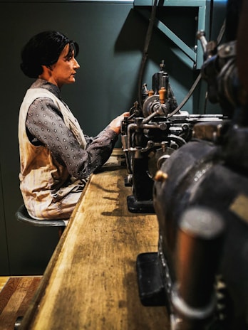 A woman is seated at a wooden workbench, focused on operating machinery, possibly in a historical or industrial setting. She wears a vintage-style blouse and vest. In the background, there is industrial equipment with intricate metal parts.