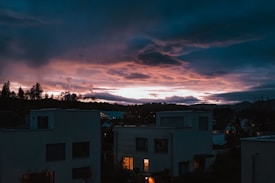 A dramatic evening sky painted with deep blues and vibrant oranges hangs over a quiet residential area. Silhouetted trees line the horizon, contrasting against the vivid colors of the sunset. Modern buildings with simple geometric shapes are visible below, some illuminated from within by warm lights, suggesting life inside.