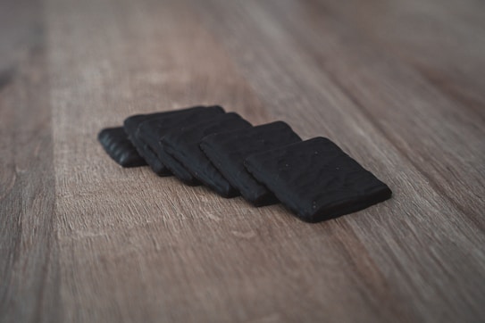 A series of dark, square objects are neatly arranged on a wooden surface. The objects have a matte finish with a slightly textured appearance, casting soft shadows on the light brown wood.