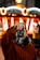 A person is holding a camera up to their face, capturing a photograph. They are wearing a brown jacket with a fur collar. In the background, there is a blurred, brightly lit carousel with circular light patterns.