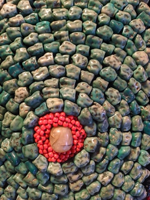 An arrangement of smooth, polished stones. Outer layers consist of bluish-green stones with irregular shapes and a speckled appearance. At the center, there is a circle of small, bright red beads surrounding a larger, smooth beige stone.