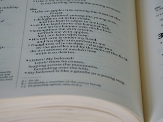 A close-up of an open book with text that appears to be poetic or biblical. The text is formatted in two columns, with numbers next to certain lines. The focus is sharp on the center of the open pages, and the pages are slightly curved at the bottom edge, indicating the book is partially open.
