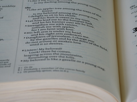 A close-up of an open book with text that appears to be poetic or biblical. The text is formatted in two columns, with numbers next to certain lines. The focus is sharp on the center of the open pages, and the pages are slightly curved at the bottom edge, indicating the book is partially open.
