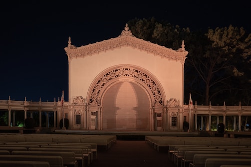 An ornate, illuminated outdoor stage with intricate architectural design, including detailed archways and finial decorations. Surrounded by symmetrical seating rows, the structure is set against a backdrop of trees at night, lit by warm artificial lighting.