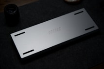A sleek, metallic rectangular object with four elongated slots and a series of small rectangular engravings down the center. The object is placed on a dark fabric surface, with a lens visible in the background.