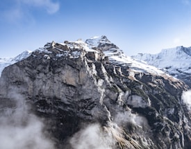 A majestic mountain range capped with snow under a clear blue sky. The rugged cliffs with layers of exposed rock create a dramatic and breathtaking landscape. Mist floats gently around the base, adding a mystical touch to the scene.